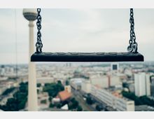 Nahaufnahme einer leeren schwarzen Schaukel, aufgehängt an Ketten, mit unscharfer Stadtlandschaft im Hintergrund. Der Berliner Fernsehturm ist prominent im linken Bildhintergrund zu sehen.