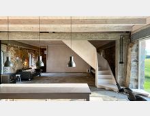 Wohnraum in einem renovierten Gebäude mit sichtbaren Backstein- und Natursteinwänden, einer freiliegenden Holzdecke und modernen Hängelampen. Eine helle Holztreppe führt nach oben, während große Fenster einen Blick auf eine grüne Außenlandschaft bieten.