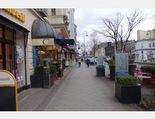Straßenszene mit Bürgersteig, flankiert von Geschäften, darunter ein Café mit Backwaren im Schaufenster. Auf der rechten Seite stehen Tische und Stühle vor mehreren Imbissständen, während vereinzelte Passanten die Straße entlanggehen, umgeben von kahlen Bäumen und einem grauen Himmel.