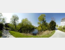 Parklandschaft mit einem kleinen See, umgeben von Bäumen mit frischem Frühlingslaub. Rechts führt ein gepflasterter Weg vorbei an einem Gebäude mit weißen Wänden und Mansardendach.