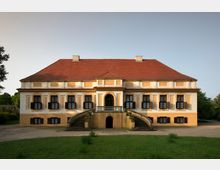 Zweigeschossiges Herrenhaus in barockem Stil mit rotem Walmdach, symmetrischer Fassade, schwarzen Fensterläden und einer zentralen Freitreppe mit geschwungenen Geländern. Das Gebäude ist von einer Wiese und vereinzelten Bäumen umgeben.