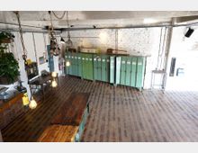 Industrieller Innenraum mit einer Wand aus weißen Ziegeln, grünen Metallspinden und hölzernem Boden. Im Vordergrund befinden sich Holztische, industrielle Lampen hängen von der Decke, und an der Seite sind verschiedene Werkstattmöbel und Pflanzen sichtbar.