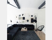 Modernes Wohnzimmer mit weiß gestrichenen Wänden, einer Collage aus gerahmten Kunstwerken, einer schwarzen Eckcouch, einem grauen Teppich und minimalistischem Mobiliar wie einem Beistelltisch und einer Stehlampe. Sichtbare Deckenträger aus Metall und ein helles Licht durch Fenster ergänzen das Design.