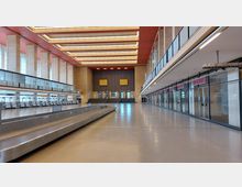 Große, lichtdurchflutete Halle mit hohen Fenstern, einer Reihe von Check-in-Schaltern auf der linken Seite und geschlossenen Geschäften mit Glasfassaden auf der rechten Seite. Eine breite Treppe führt am Ende der Halle zu mehreren Türen, die von einer monumentalen Wand eingerahmt sind.