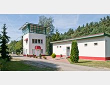 Ein rechteckiges Gebäude mit flachem Dach und ein schmaler, mehrstöckiger Turm mit grüner Fensterstruktur stehen nebeneinander in einer parkähnlichen Umgebung mit Bäumen und Gehweg. Beide Gebäude sind in Beige und Rot gehalten, mit einem Vordach über dem Eingang des Turms.