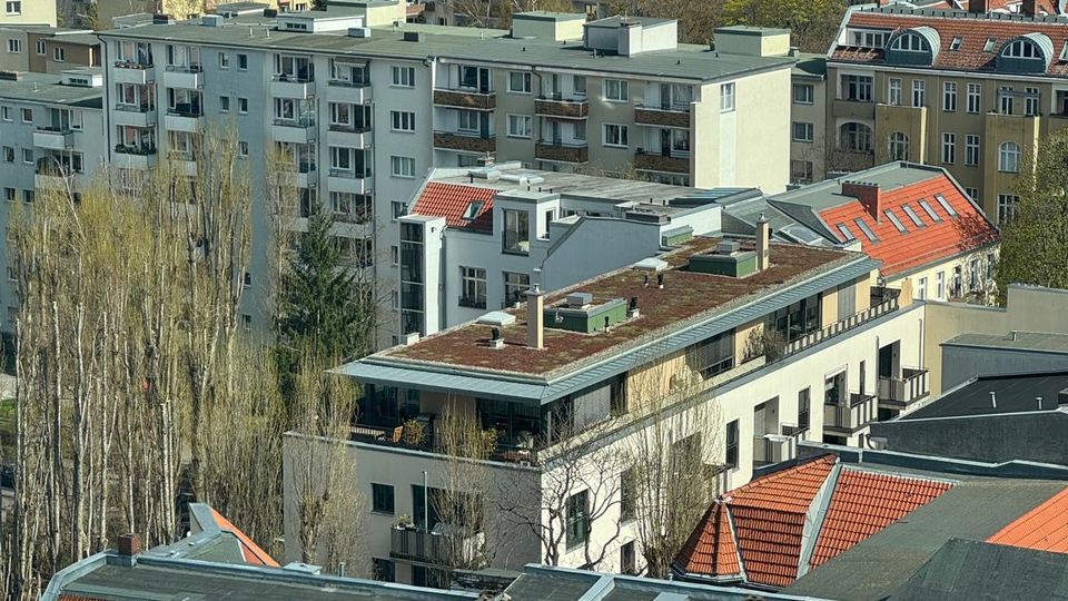 Blick über ein dicht bebautes Stadtviertel mit mehrstöckigen Wohnhäusern, Flachdächern und roten Ziegeldächern. Im Hintergrund sind weitere Wohnblöcke und höhere Büro- bzw. Hochhausgebäude am Stadtrand zu sehen, dazwischen einzelne Baumreihen.