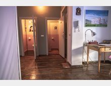 Ein Innenraum mit dunklem Holzboden, zwei leicht geöffnete Türen zu Toilettenräumen mit rosafarben-weißen Wänden, und einem einfachen Holztisch rechts im Bild, auf dem eine Schreibtischlampe und Malutensilien stehen. An der Wand hängen ein Bild eines Hochhauses und ein kleiner religiöser Wandanhänger.