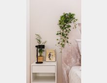 Nachttisch mit weißer Oberfläche, darauf eine schwarze Lampe, eine kleine grüne Pflanze in einem Topf und ein gerahmtes Bild. Daneben eine Wand mit hängendem Efeu und ein Bett mit einer weißen Steppdecke, teilweise sichtbar.