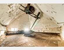 Ein verlassenes Industriegebäude mit einem großen Raum unter einem gewölbten Dach. Sichtbar sind eine rostige Maschinerie, abblätternder Putz an den Wänden und mehrere kleine vergitterte Fenster, die natürliches Licht hereinlassen.