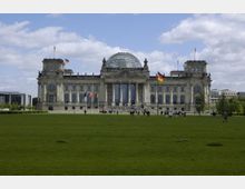 Frontale Ansicht des Reichstagsgebäudes in Berlin, mit seiner markanten Glaskuppel und klassizistischer Steinfassade. Im Vordergrund befindet sich eine große Rasenfläche mit verstreuten Besuchern, während mehrere Fahnen, darunter die deutsche Flagge, das Gebäude umgeben.