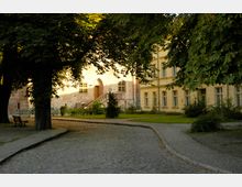 Historischer Hof mit Kopfsteinpflasterwegen, umgeben von Bäumen und Grünflächen; im Hintergrund ein Gebäude aus gelbem Putz mit großen Fenstern sowie ein Backsteingebäude mit Rundbogenportal und Treppe.