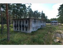 Verfallenes zweistöckiges Gebäude mit beschädigten Fenstern und Rußspuren an der Fassade, umgeben von hohem Gras und Kiefernwäldern; im Hintergrund ein weiteres ähnliches Gebäude unter klarem Himmel.