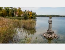 Eine Steinfigur einer Frau mit Blumenkranz, die auf einem kleinen Steinhaufen im Wasser eines ruhigen Sees steht; im Hintergrund ein Ufer mit Schilfrohr, Bäumen in herbstlichen Farben und einem Gebäude mit rotem Dach.