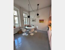 Heller Essbereich mit grauem Boden, einem runden weißen Tisch und vier weißen Stühlen mit braunen Sitzflächen. Im Raum befinden sich große Fenster, eine Hängelampe mit schwarzen Leuchten und eine pastellrosa Küchenzeile an der rechten Wand.