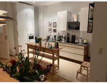 Eine moderne Küche mit weißen Hochglanzschränken, einer Arbeitsplatte aus Edelstahl, einer Dunstabzugshaube und verschiedenen Küchengeräten. In der Mitte steht ein hölzerner Küchenwagen, im Vordergrund ein Blumenstrauß.