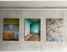 Drei gerahmte Fotografien hängen an einer grauen Betonwand. Die Bilder zeigen Innenaufnahmen: links eine Küchenzeile mit Fliesen in Gelb-, Orange- und Weißtönen, in der Mitte ein grün gestrichener Raum mit Heizkörper und Fliesenmuster, rechts ein florales Muster mit großen Blüten in Pastellfarben.