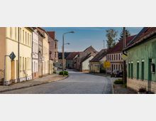 Straßenszene in einer kleinen Stadt mit Kopfsteinpflaster, umgeben von zweigeschossigen, überwiegend hell gestrichenen Wohnhäusern. Verkehrsschilder, Laternenmasten und ein gelbes Ortshinweisschild sind entlang der Straße sichtbar.