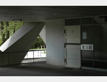 Unter einem Betonvordach befindet sich der Eingangsbereich eines Gebäudes mit einer weißen Tür und der Aufschrift „8“. Links stützt eine schräge Betonsäule das Dach ab, im Hintergrund sind Bäume und eine asphaltierte Fläche mit rot-weißen Pollern sichtbar.