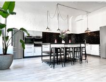 Moderne Küche mit weiß gestrichenen Wänden und dunklen Fliesen im U-Bahn-Stil an der Rückwand. Zentral steht ein großer Küchenblock mit schwarzen Barstühlen, darüber hängen mehrere industrielle Hängelampen; links ein großer Blumentopf mit einer Pflanze.