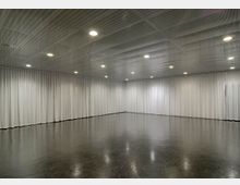 Großer, leerer Raum mit glänzendem dunklem Boden, weißen, deckenhohen Vorhängen an den Wänden und einer perforierten Decke mit mehreren eingelassenen runden Deckenleuchten.