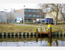 Industriegebiet mit Gebäuden aus Metall und Glas, einem Zaun, einem Baum und einem Wasserkanal im Vordergrund. Ein Geländer führt zu Stufen, die auf eine kleine Anlegestelle mit gelbem Poller führen.