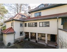 Großes Wohnhaus mit cremefarbener Fassade und einem roten Ziegeldach, das mehrere Dachgauben und Dachfenster aufweist. Im Erdgeschoss befindet sich eine überdachte Terrasse mit massiven Säulen, Gitterfenstern und einer dunkelgrünen Eingangstür; umgeben von einem gepflasterten Hof und Bäumen im Hintergrund.