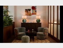 Gemütliche Sitzecke in einem eleganten Raum mit zwei gepolsterten Sesseln und einem kleinen runden Tisch auf einem gemusterten Teppich. Im Hintergrund steht eine dunkle Kommode mit zwei Tischlampen und Wanddekor, umrahmt von Pflanzen und einer Holzwand mit Glasverkleidung.