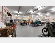Ausstellungsraum in einem Museum mit historischen Fahrzeugen, darunter ein cremefarbenes und ein grünes Oldtimer-Auto, Fahrrädern und Motorrädern. Die Wände sind mit historischen Fotografien und Tafeln dekoriert, die Informationen zu den Exponaten vermitteln.