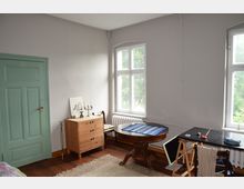 Ein helles Zimmer mit weißen Wänden, einem Holzboden und zwei großen Fenstern, die Tageslicht hineinlassen. Es enthält eine grüne Holztür, eine Holzanrichte mit Kerzenleuchter, einen runden Holztisch mit blauer Tischdecke und einen schlichten Arbeitstisch auf Böcken.