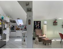 Offene Wohnküche und Wohnzimmer in einem Dachgeschoss mit schrägen Decken. Links befindet sich eine weiße Küche mit farbenfrohen Fliesen und einem Dachfenster, rechts ein Wohnzimmer mit grün gestrichener Wand, einem Sessel, einer Stehlampe und Dekorationen.