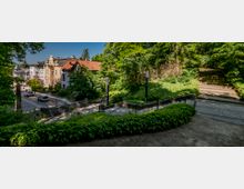 Treppenanlage in einer grünen Umgebung mit üppiger Vegetation, flankiert von dekorativen Laternen. Im Hintergrund sind gepflasterte Straßen und historische Wohnhäuser mit verzierten Fassaden sichtbar.