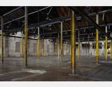 Großer, leerer Industrie-Raum mit hohen, gelben Stahlpfeilern, einer offenen Dachkonstruktion aus Metall und Holz sowie freiliegenden Backsteinwänden. Der Boden besteht aus glattem Beton, und die Umgebung wirkt wie eine umgebaute Fabrikhalle oder Lagerhalle.