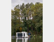 Ein modernes, weißes Hausboot mit großen Fenstern liegt auf einem ruhigen Gewässer, umgeben von einem Ufer mit hohen Bäumen und dichter Vegetation. Im Hintergrund stehen kleine Holzhütten zwischen den Bäumen.