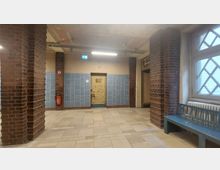 Ein Flur in einem älteren Gebäude mit Backsteinpfeilern und hellen Bodenfliesen. Die Wände sind teilweise mit blauen Kacheln verkleidet, eine Holztür mit abgeblätterter Farbe ist in der Mitte sichtbar, daneben ein Feuerlöscher und ein Fenster mit Gittermuster.