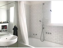 Ein modernes Badezimmer mit einer Badewanne und einer weißen Fliesenwand. Auf der rechten Seite befindet sich eine Dusche mit abnehmbarem Brausekopf, daneben ein Fenster; links ein Waschbecken mit Spiegel und Ablage.