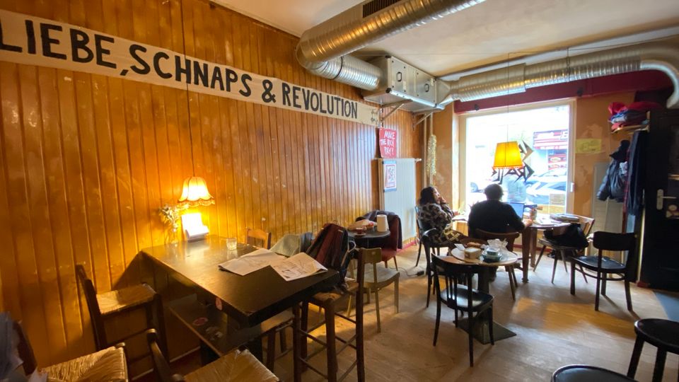 Gemütlicher Café- oder Barinnenraum mit Holzvertäfelung, gemischten Tischen und Stühlen sowie einem großen Wandbanner „LIEBE, SCHNAPS & REVOLUTION“. Sichtbare Lüftungsrohre an der Decke und ein großes Fenster zur Straße sorgen für industriellen Charakter und Tageslicht.