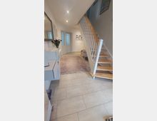 Heller Eingangsbereich mit beiger Fliesenboden, einer Holztreppe mit weißen Geländern, sowie einem Lilateppich und einer kleinen Sitzbank. An der linken Wand befinden sich hängende Kommoden und ein Wandspiegel, geradeaus sind weiße Doppeltüren sichtbar.