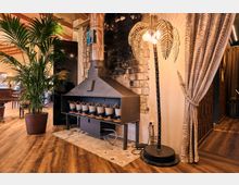 Warmly lit restaurant interior with wooden floors, a large black metal fireplace hood and chimney against a dark brick wall, and a potted palm beside it. A palm-shaped floor lamp and patterned curtain partition define a cozy corner of the space.