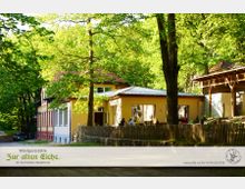 Waldgaststätte mit gelber Fassade und rotem Dach, umgeben von dichtem, grünem Laubwald. Im Vordergrund befindet sich ein kleiner eingezäunter Außenbereich mit Tischen und Stühlen unter Bäumen.