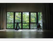 Ein heller Raum mit großen bodentiefen Fenstern, die den Blick auf grüne Bäume freigeben. Zwei Personen machen auf Holzfußboden Yoga- oder Dehnübungen, das Licht fällt durch die Fenster auf den Boden.