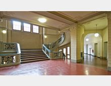 Treppenhaus eines historischen Gebäudes mit geschwungener Treppe, verziertem Geländer und roten Marmorböden. Der Raum umfasst hohe Fenster, verzierte Stuckdecken und Rundbogenöffnungen, die in angrenzende Flure führen.