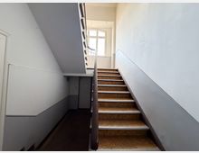 Innenansicht eines Treppenhauses mit gerader Treppe, braun gemusterten Stufen und dunkelbraunem Handlauf, die zu einem Podest führt. Helle Wände mit grauem Sockelbereich und ein großes Fenster am Treppenabsatz lassen Tageslicht in den Raum.