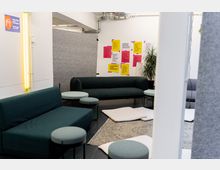 Ein moderner Innenraum mit dunkelgrünen Sofas, runden Hockern und weißen Sitzkissen auf Teppichen. An der weißen Wand hängen bunte Plakate mit Text, daneben befindet sich eine Pflanze; ein gelbes Neonlicht ist an einer Seitenwand angebracht.