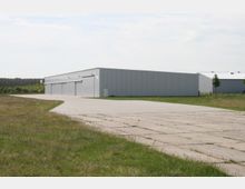 Ein großes, graues Industriewerkstattgebäude mit flachem Dach und Rolltoren steht inmitten einer offenen Grünfläche. Der gepflasterte Weg führt am Gebäude entlang, und im Hintergrund sind weitere ähnliche Gebäudestrukturen sowie Bäume sichtbar.