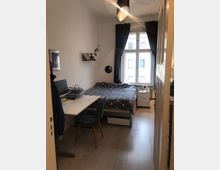 Kleines Schlafzimmer mit einem Einzelbett, das an einer Wand mit einem Regal steht. Ein Schreibtisch mit einem Stuhl befindet sich gegenüber dem Bett, und ein Fenster mit dunklen Vorhängen lässt Tageslicht in den Raum.