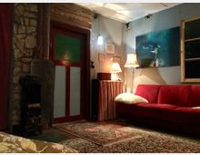 Ein gemütlich eingerichtetes Wohnzimmer mit einer roten Couch, einem gemusterten Teppich und einem kleinen Ofen aus Metall an einer Wand mit heller Steinverkleidung. Das Zimmer enthält außerdem eine Stehlampe mit Stoffschirm, eine Holztür mit rotem Rahmen sowie eine Wanddekoration in Form von Gemälden und Drucken.