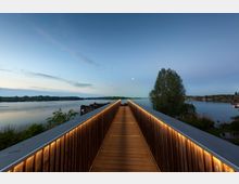 Ein beleuchteter Holzsteg führt über das Wasser eines ruhigen Sees, umgeben von Bäumen und einer flachen Uferlandschaft; der Himmel ist klar mit Mondlicht und Abendstimmung.