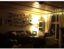 Wohnzimmer mit Holzbalkendecke, einer Sitzecke mit Sofa und Sessel, einer Wand mit zahlreichen gerahmten Bildern sowie einem Fenster mit langen weißen Vorhängen. Im Raum stehen eine kleine Lampe und ein Schrank, der Beleuchtung und Dekoration ergänzt.