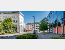 Straßenszene in einer modernen Stadt mit mehrstöckigen Wohn- und Bürogebäuden; links ein weißes Gebäude mit großen Fenstern, rechts ein Gebäude mit einer Fassade aus Glas und roten Elementen. Die Straße in der Mitte ist von Bäumen gesäumt, mit geparkten Autos und Fahrrädern im Vordergrund.