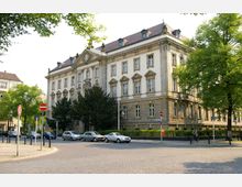 Ein historisches, mehrstöckiges Gebäude mit symmetrischer Fassade im neoklassizistischen Stil, verziert mit Säulen und flachen Erkern, steht an einer Straßenecke. Im Vordergrund sind parkende Autos, Verkehrsschilder und grüne Bäume sichtbar.